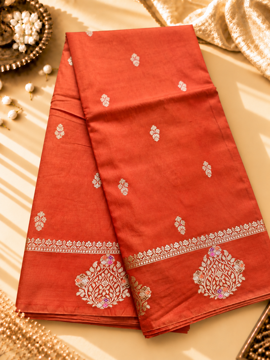A vibrant Coral Orange Banarasi viscose silk saree featuring delicate traditional butis, intricate floral motifs and elegant Minakari weaving. The warm coral-orange base beautifully complements the metallic and colourful detailing,