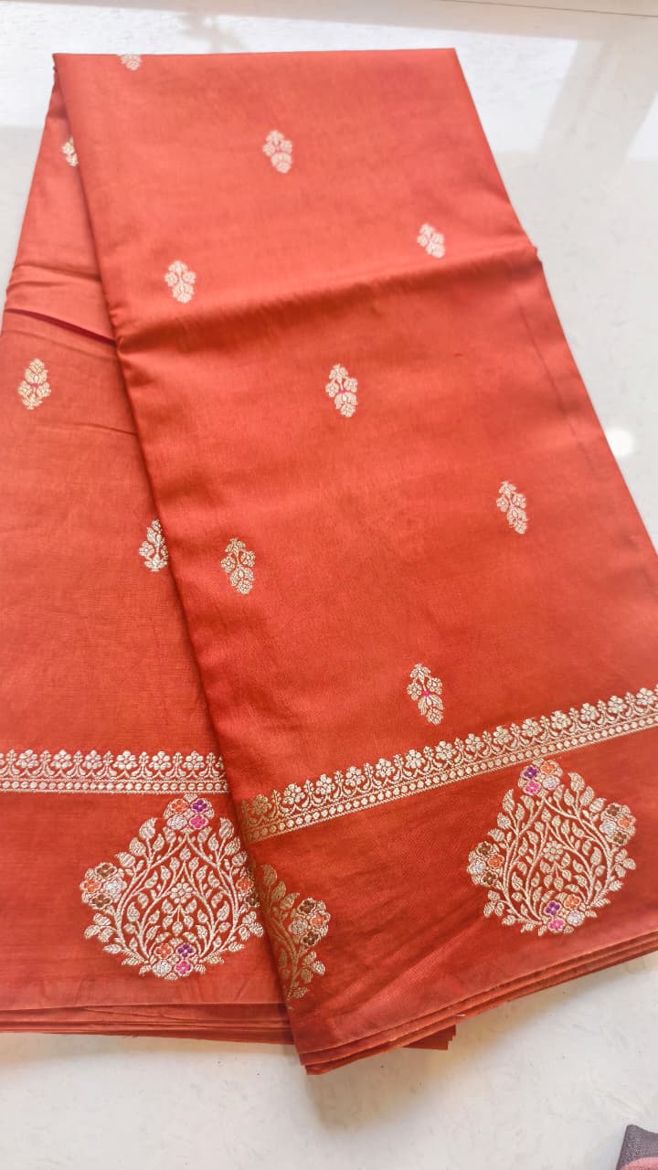 A vibrant Coral Orange Banarasi viscose silk saree featuring delicate traditional butis, intricate floral motifs and elegant Minakari weaving. The warm coral-orange base beautifully complements the metallic and colourful detailing,