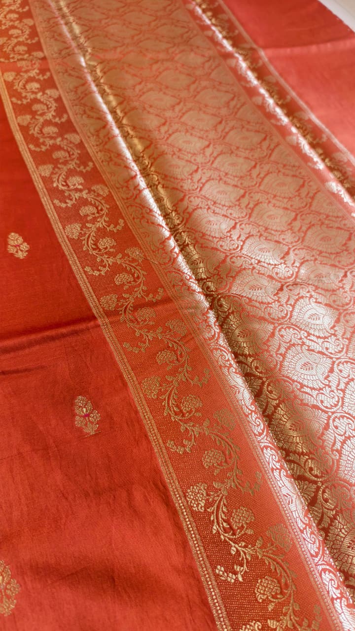 A vibrant Coral Orange Banarasi viscose silk saree featuring delicate traditional butis, intricate floral motifs and elegant Minakari weaving. The warm coral-orange base beautifully complements the metallic and colourful detailing,
