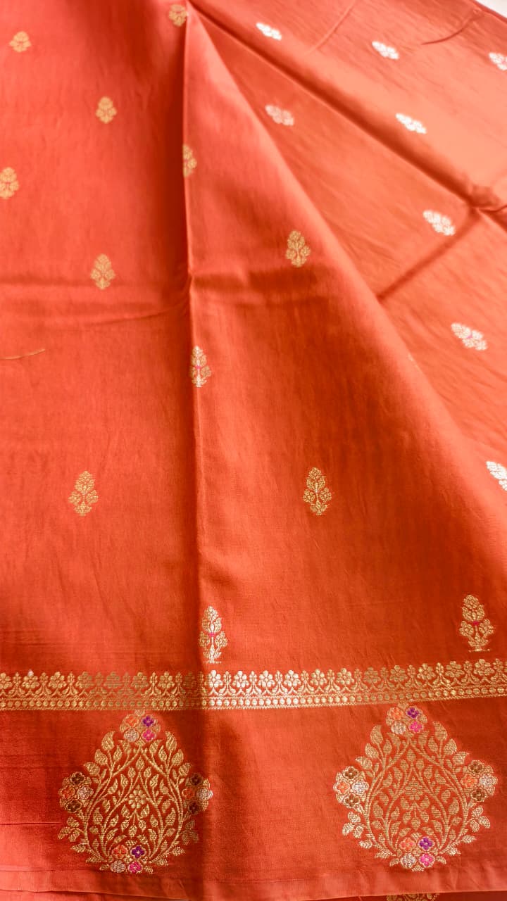 A vibrant Coral Orange Banarasi viscose silk saree featuring delicate traditional butis, intricate floral motifs and elegant Minakari weaving. The warm coral-orange base beautifully complements the metallic and colourful detailing,