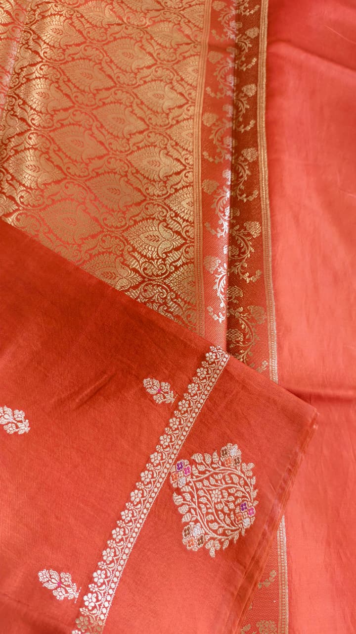 A vibrant Coral Orange Banarasi viscose silk saree featuring delicate traditional butis, intricate floral motifs and elegant Minakari weaving. The warm coral-orange base beautifully complements the metallic and colourful detailing,